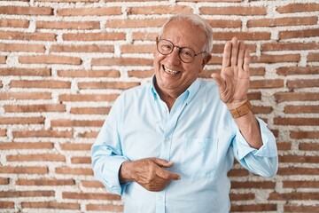 Senior man with grey hair standing over bricks wall waiving saying hello happy and smiling, friendly welcome gesture © Krakenimages.com