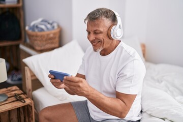 Middle age grey-haired man watching video on smartphone sitting on bed at bedroom