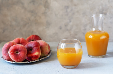 Peach juice in a glass and pitcher with raw fresh flate peaches on silver tray. Grey concrete background with copy space