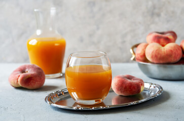 Peach juice in a glass on silver tray, pitcher with juice, ripe fresh flate peaches. Grey concrete background with copy space