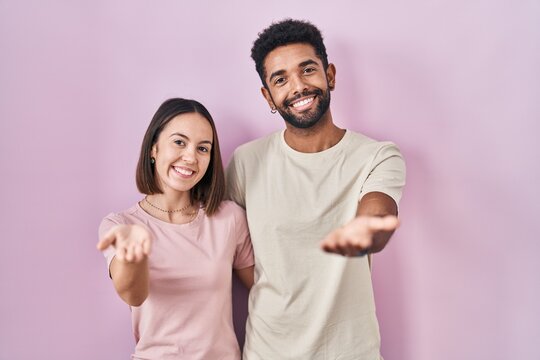 Young hispanic couple together over pink background smiling cheerful offering palm hand giving assistance and acceptance.