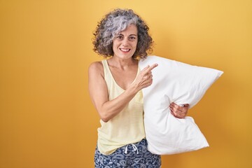 Middle age woman with grey hair wearing pijama hugging pillow cheerful with a smile on face pointing with hand and finger up to the side with happy and natural expression