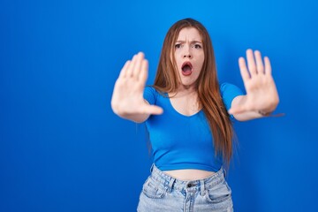 Redhead woman standing over blue background doing stop gesture with hands palms, angry and...