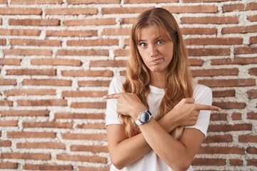 Young caucasian woman standing over bricks wall pointing to both sides with fingers, different direction disagree