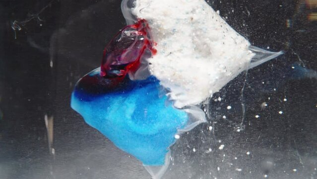 A close-up shot of a soluble packet for a dishwasher dissolving underwater. The red gel and blue-white powder are slowly releasing and dispersing in the water.