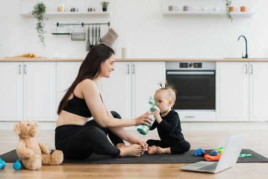 Beautiful adult female and child sitting cross-legged on yoga mat and drinking from sports bottle in kitchen. Energetic mother and daughter restoring water balance after effective home workout.