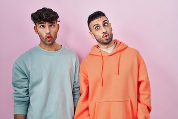 Young hispanic gay couple standing over pink background making fish face with lips, crazy and comical gesture. funny expression.