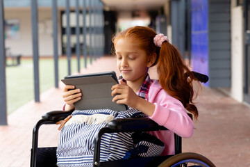 Happy biracial schoolgirl in wheelchair, using tablet in school corridor