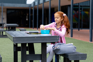Happy biracial schoolgirl sitting at table with packed lunch in schoolyard