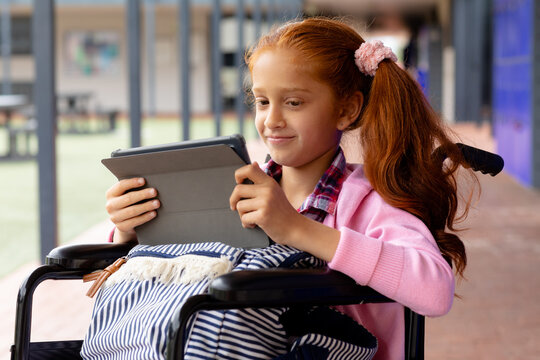 Happy biracial schoolgirl in wheelchair, using tablet in school corridor