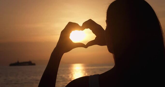 Happy woman looking at sea sunset, showing heart gesture with fingers. Rear view silhouette woman at sunset, close up making heart figure with hands holding inside the sun. Summer dream.