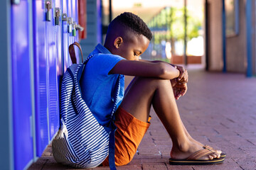 Sad african american schoolboy with school bag sitting by lockers in school, with copy space