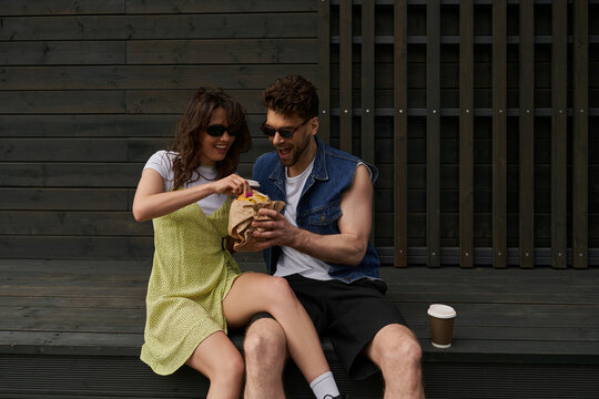 Cheerful and trendy romantic couple in sunglasses and summer outfits holding fresh bun while sitting near coffee to go and wooden house at background, countryside exploration concept