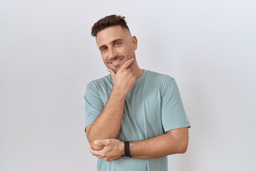 Fototapeta premium Hispanic man with beard standing over white background looking confident at the camera smiling with crossed arms and hand raised on chin. thinking positive.
