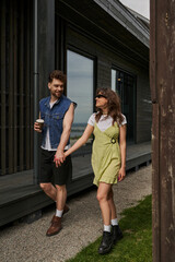 Obraz premium Smiling brunette woman in sunglasses and sundress holding hand of bearded boyfriend with takeaway coffee and walking together near wooden houses in rural setting, outdoor enjoyment concept