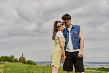 Stylish bearded man in denim vest touching hand of romantic girlfriend in sunglasses and sundress while standing together with rural setting at background, countryside retreat concept