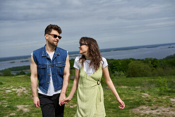 Cheerful brunette woman in sunglasses and stylish sundress holding hand and looking at boyfriend in denim vest and standing on grassy hill, countryside wanderlust and love concept, tranquility