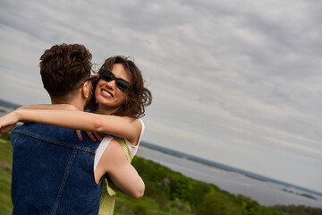 Smiling brunette woman in summer outfit and sunglasses embracing stylish boyfriend in denim vest with rural landscape and cloudy sky at background, love story and countryside adventure