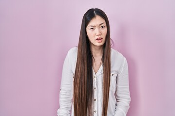 Chinese young woman standing over pink background in shock face, looking skeptical and sarcastic, surprised with open mouth