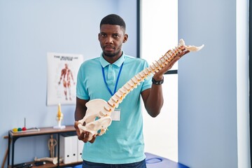 African american man holding anatomical model of spinal column relaxed with serious expression on...