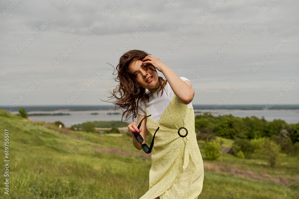 Wall mural Positive brunette woman in trendy sundress holding sunglasses and touching hair while standing and spending time with blurred natural landscape and cloudy sky at background, summertime joy