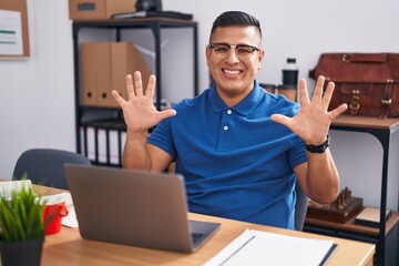 Young hispanic man working at the office with laptop showing and pointing up with fingers number ten while smiling confident and happy.