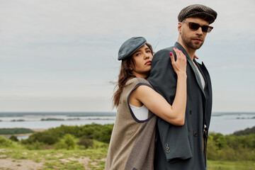 Portrait of brunette woman in newsboy cap and vest embracing bearded boyfriend in sunglasses and jacket while standing with blurred landscape at background, trendy twosome in rustic setting