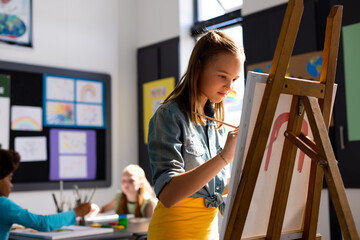 Obraz premium Happy caucasian schoolgirl painting using brush and easel in school art class