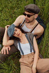 Naklejka na ściany i meble Top view of fashionable brunette woman in newsboy cap and vintage outfit relaxing near bearded boyfriend in sunglasses on grassy meadow, fashionable couple surrounded by nature, romantic getaway