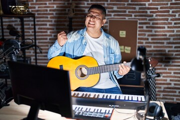 Hispanic young man playing classic guitar at music studio very happy and excited doing winner gesture with arms raised, smiling and screaming for success. celebration concept.