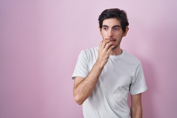 Young hispanic man standing over pink background looking stressed and nervous with hands on mouth biting nails. anxiety problem.