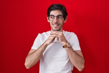 Young hispanic man standing over red background laughing nervous and excited with hands on chin looking to the side