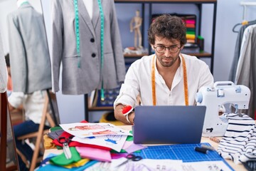 Young hispanic man tailor using laptop at clothing factory