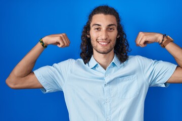 Naklejka premium Young hispanic man standing over blue background showing arms muscles smiling proud. fitness concept.