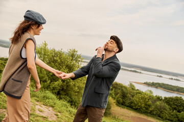 Fototapeta na wymiar Fashionable man in newsboy cap and jacket showing secret gesture and holding hand of cheerful girlfriend while standing with nature at background, fashion-forwards in countryside