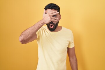 Hispanic man with beard standing over yellow background peeking in shock covering face and eyes with hand, looking through fingers with embarrassed expression.