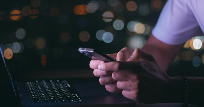 Close-up of male hands hold mobile phone, texting message while working on a laptop computer on background of a night city. Man freelancer using a mobile phone while working at computer in the evening