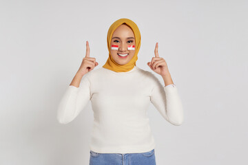 Smiling young Asian Muslim woman wearing hijab pointing her finger up, showing a place for your...
