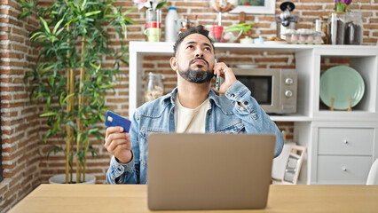 Young latin man arguing on smartphone holding credit card at dinning room