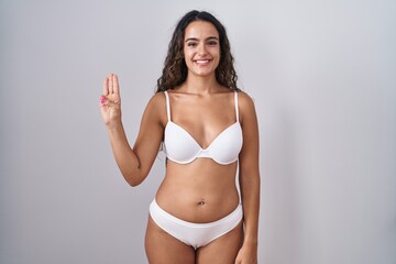 Fototapeta premium Young hispanic woman wearing white lingerie showing and pointing up with fingers number three while smiling confident and happy.