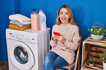Young woman using smartphone waiting for washing machine at laundry room
