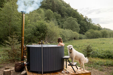 Woman bathing in outdoor hot tub while resting with her cute dog at house in mountains. Concept of...
