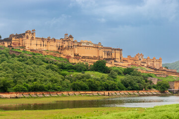 Amer fort palace with beautiful domes, arches, and balconies stands majestically amid a blue cloudy sky on the top of the mountain located near the lake surrounded by green grassland. Rajasthan, India - obrazy, fototapety, plakaty