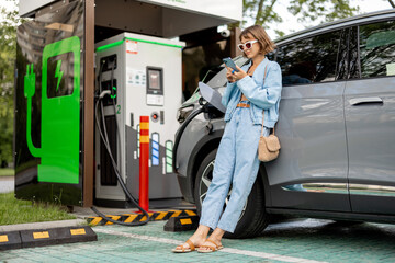 Young woman using a smart phone, while waiting for her electric car to charge on a public charging...