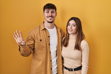 Young hispanic couple standing over yellow background showing and pointing up with fingers number four while smiling confident and happy.