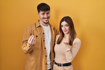 Young hispanic couple standing over yellow background doing money gesture with hands, asking for...