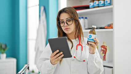 Obraz na płótnie Canvas Young beautiful hispanic woman doctor using touchpad holding medication bottle thinking at clinic