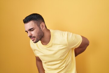 Young hispanic man standing over yellow background suffering of backache, touching back with hand, muscular pain