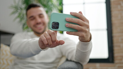 Young hispanic man watching video on smartphone sitting on sofa at home
