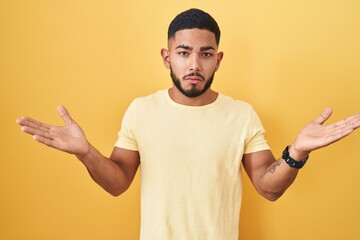 Young hispanic man standing over yellow background clueless and confused expression with arms and hands raised. doubt concept.